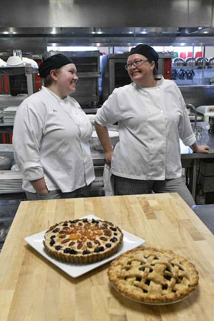 Brittani Crane and Ellie Black, wearing white chef's aprons, smile at one another. Two pies sit on a counter in front of them.