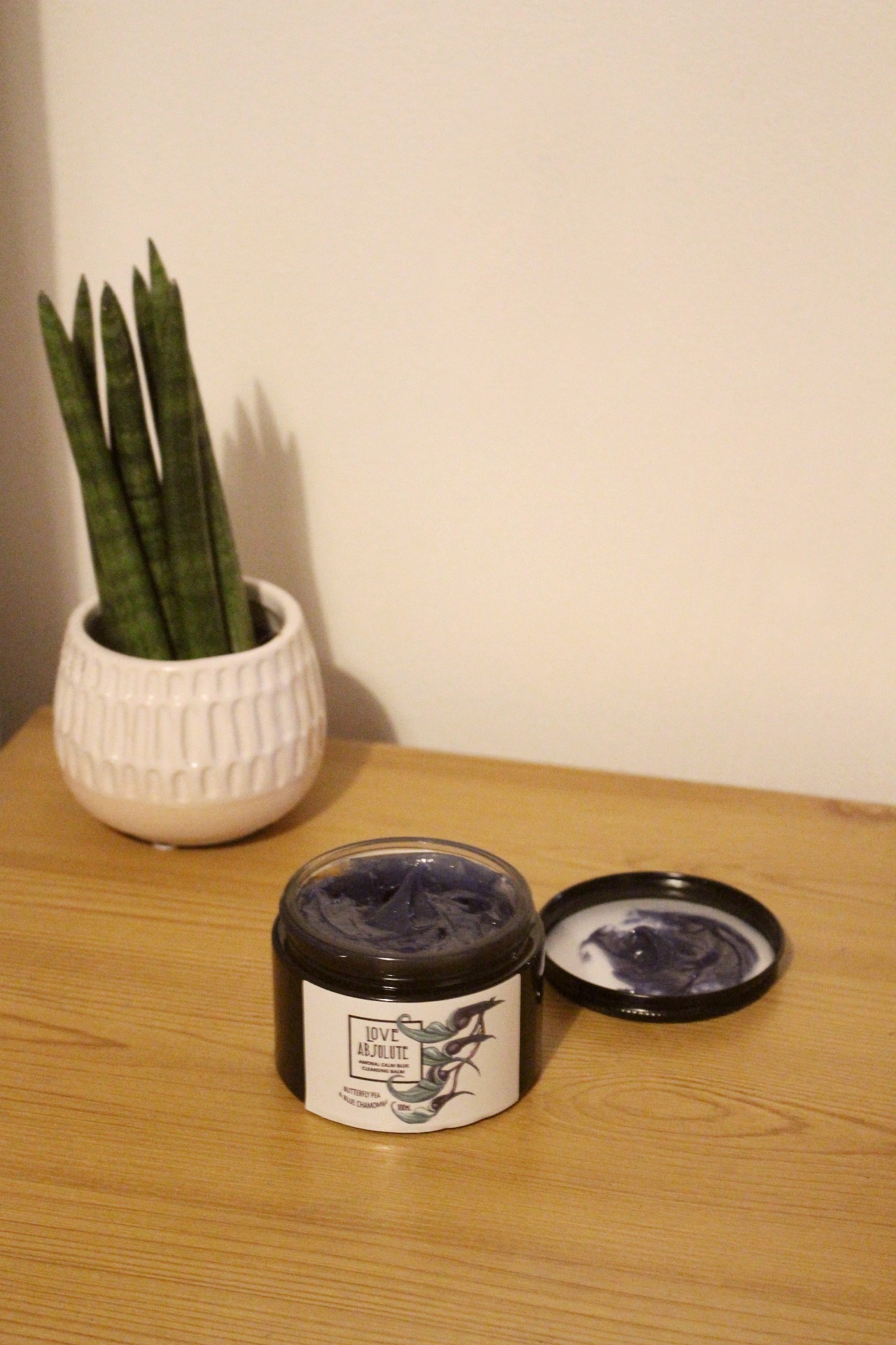 A dark coloured jar sat on a brown wooden surface. The jar has its lid removed and you can see a blue-purple product inside. The label on the jar reads "Love Absolute Amoral Calm Blue Cleansing Balm". In the background is a green succulent plant in a white pot.