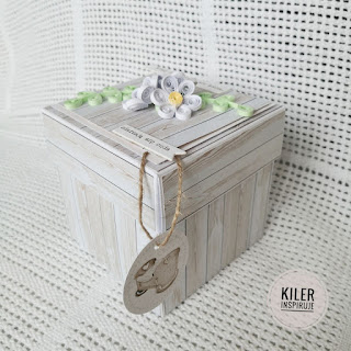 Ekspldujące pudełko ozdobione papierem z motywem desek i białymi kwiatami na wieczku.