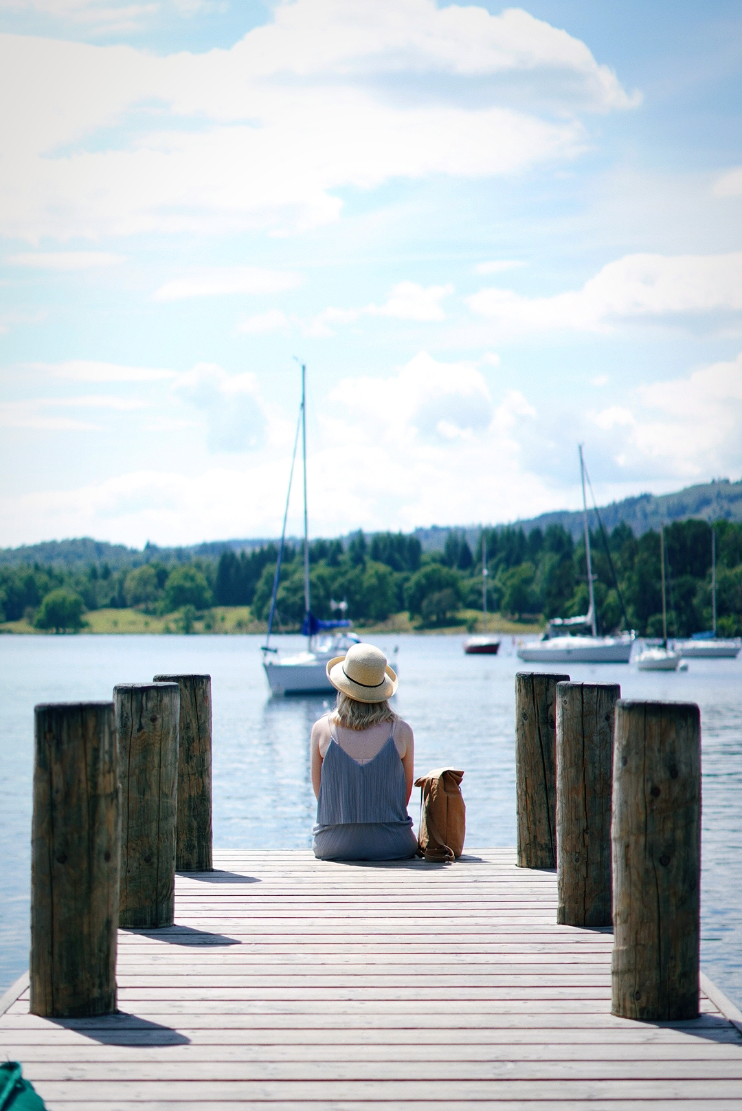 a wooden jetty overlooking a boating lake, a girl in a sun hat is sat at the end with her legs dangling over the edge