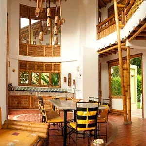 A imagem mostra uma sala circular com teto alto, grande janelas em madeira tipo bamboo, no centro uma mesa quadrada em mámore branco com 4 cadeiras e um lustre com cinco luminárias.