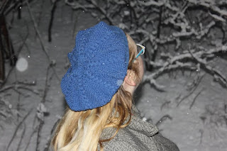 A blue tam with braid cables and moss stitch on a blonde woman looking up at a tall hedgerow as snow falls.
