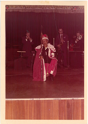 Charlie dressed in a royal robe and crown, and is holding a spear decorated with insignia of a tribe, as opposed to a sceptre similar to the Crown Jewels. He is seated, and looks quite bashful! There are people playing trumpets behind him