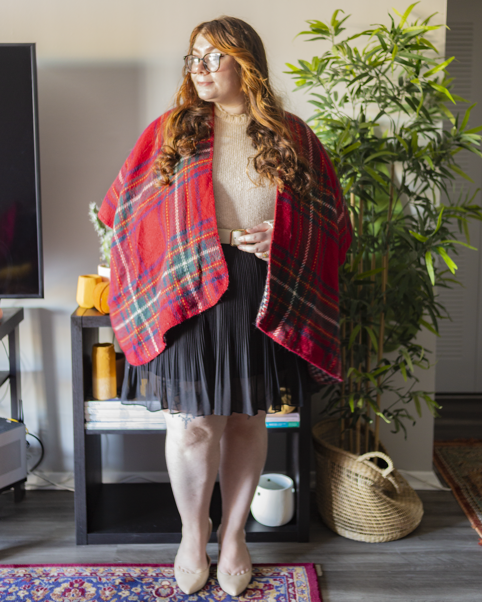 An outfit consisting of a red, green, white and blue plaid poncho over a cream sweater with a lace collar tucked into a black pleated mini skirt, black tights and black ankle boots.