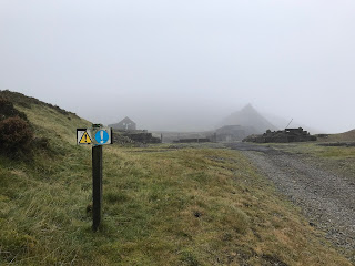 A photograph of a small warning sign with the ruined buildings and slag heap of the long abandoned New Glencrieff Mine in the distance.  Photograph by Kevin Nosferatu for the Skulferatu Project.