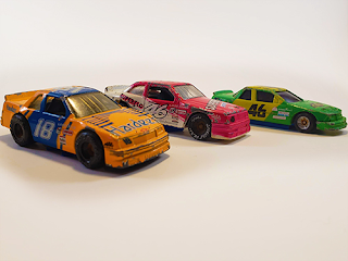 Three stock car toy cars. The one closest to the camera is orange and blue, number 18, sponsored by Hardees. The car in the middle is pink and white, number 46, sponsored by Superflo. The final car is green and yellow, number 46, sponsored by Chevy