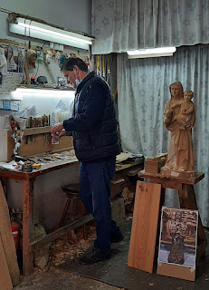 homem em bancada de trabalho ao lado de uma imagens de uma santa em madeira com algumas ferramentas e madeira