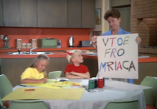 Alice, Cindy, and Jan make posters for Marcia in the kitchen. Cindy's says Vtoe Fro Mriaca.