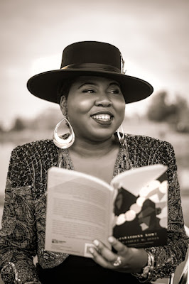 Black and white photo of Ashley M Jones posing and holding her book REPARATIONS NOW! while she stares off into the distance.