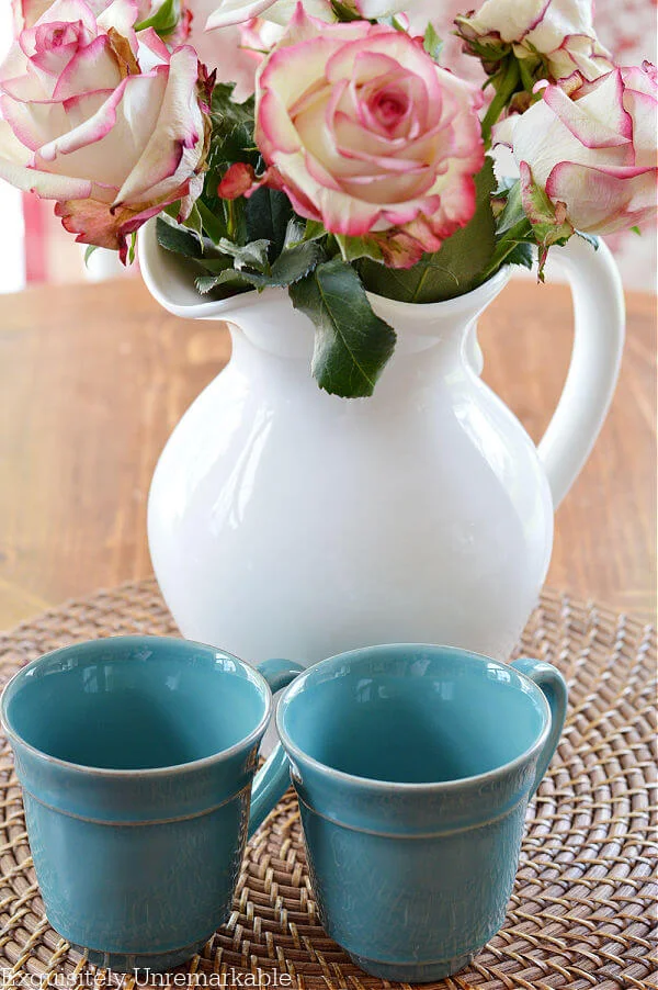 Two teal mugs near a pitcher of pink roses