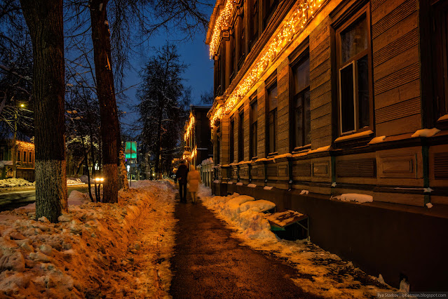 Пешеходы идут по дорожке вдоль дома с новогодней подсветкой