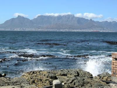 ID: waves of water crash against large rocks along the shore, with sharp shadows from the sun shining in the blue sky and Table Mountain in the background.