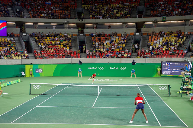 Kristen Flipkens saca durante partida contra Venus Williams na Rio 2016. Imagem em plano aberto, aparecendo toda a quadra e parte da arquibancada.