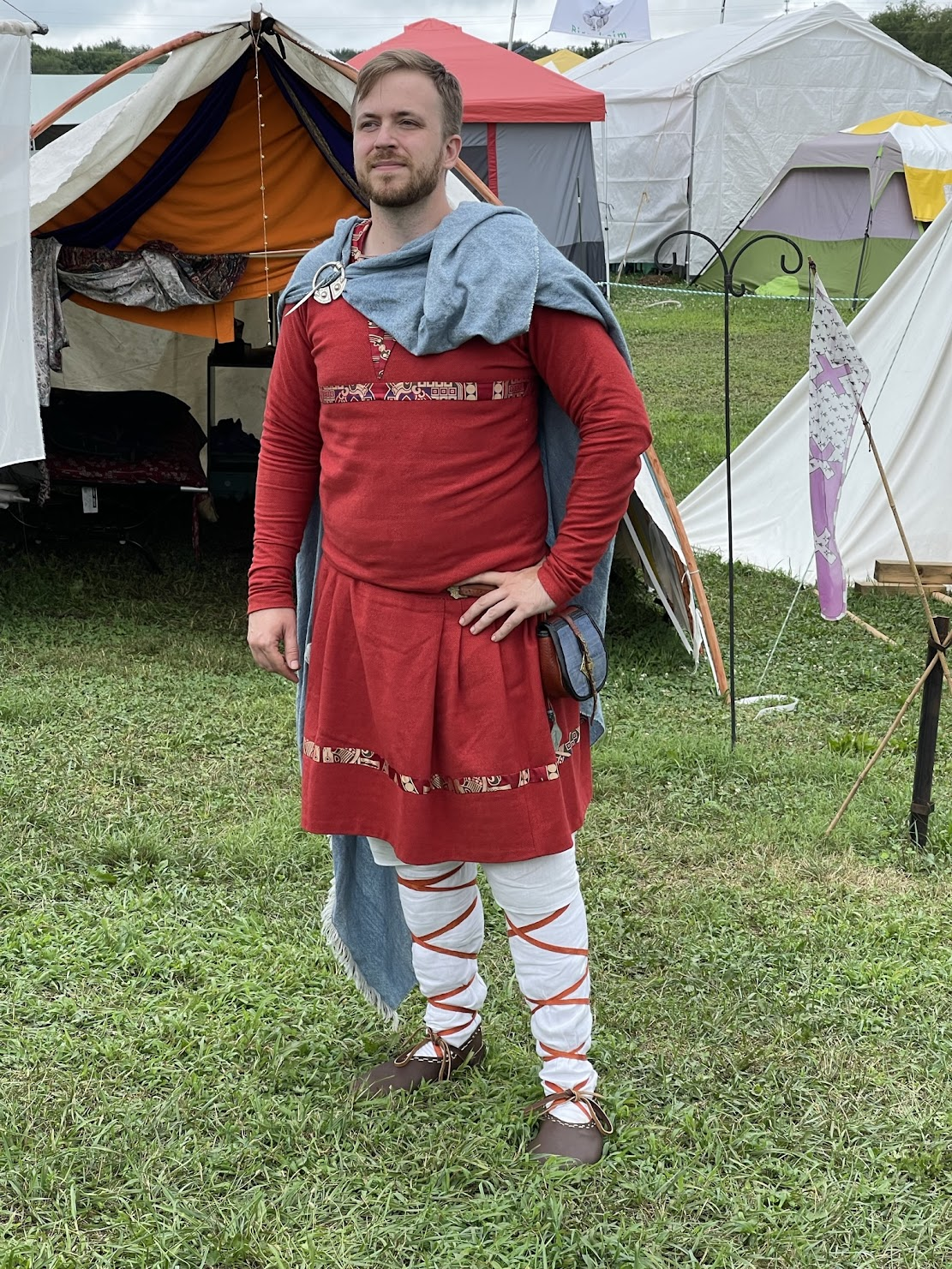 Alec standing in the outfit described in this post, with a bright red tunic with silk trimmings, a blue cloak, and white hose with red cross-garters