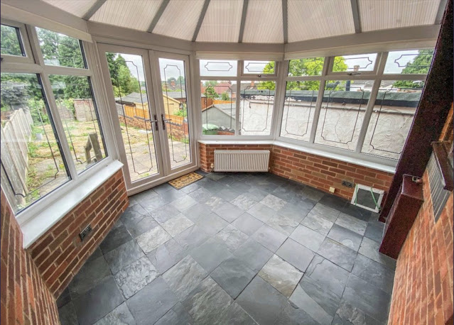 photo taken from doorway looking into conservatory. It's got a dark grey tiled floor and exposed brick walls with 4 walls taken up with large windows and french doors out to the garden. The roof has translucent panels which reach to a point in the ceiling.