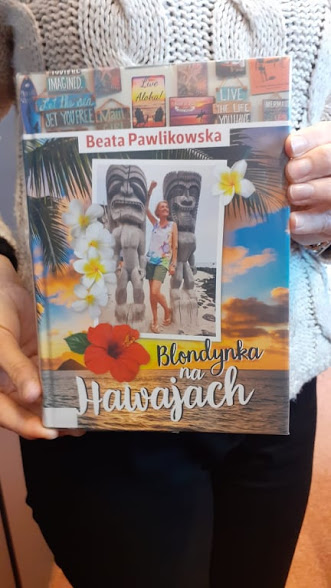 Pani w białym sweterku i czarnych spodniach prezentuje książkę Beaty Pawlikowskiej pod tytułem Blondynka na Hawajach.