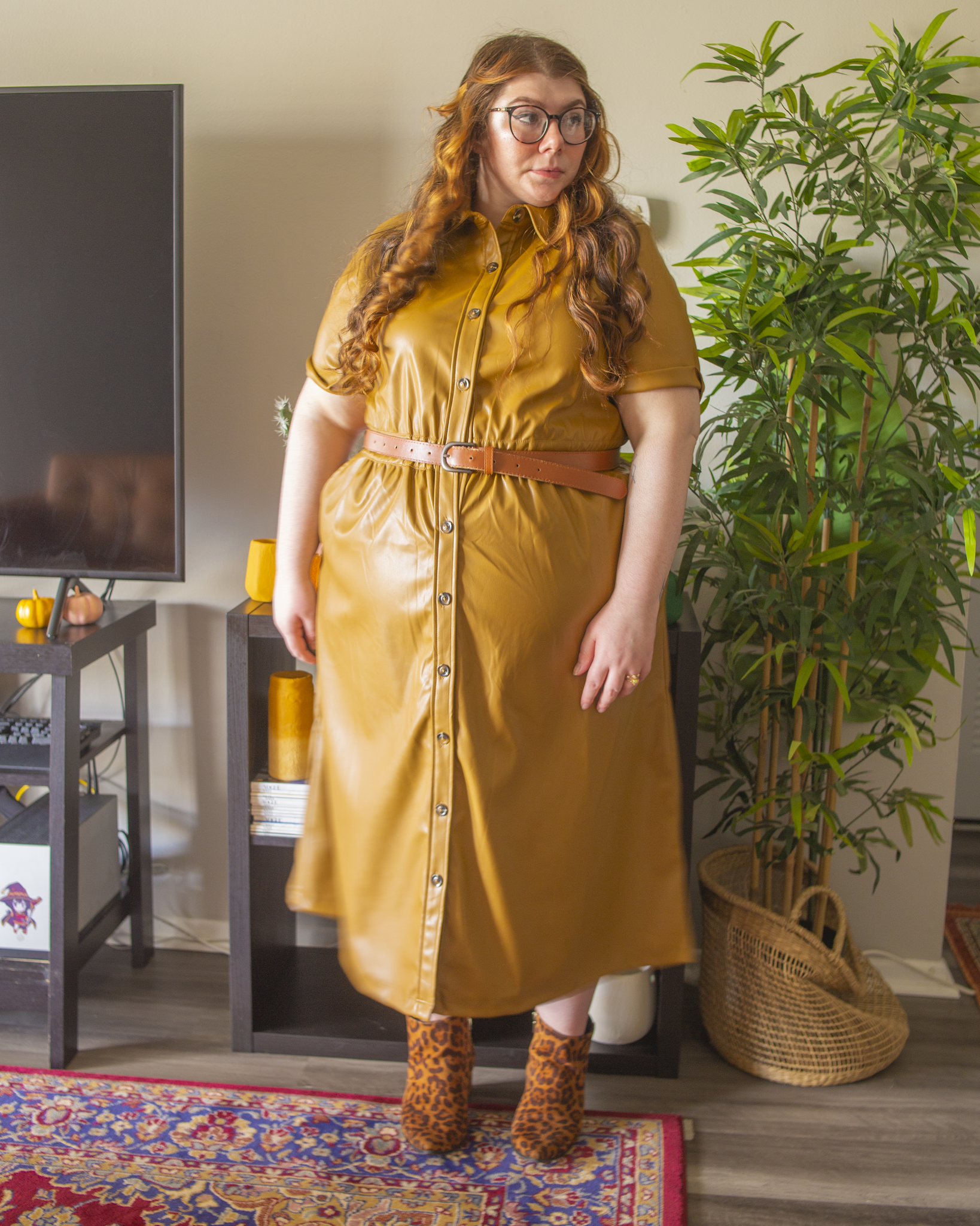 An outfit consisting of a wide brim camel panama hat, an olive green brown short sleeve faux leather shirt midi dress, belted with a dark brown belt, and black on brown animal print boots.