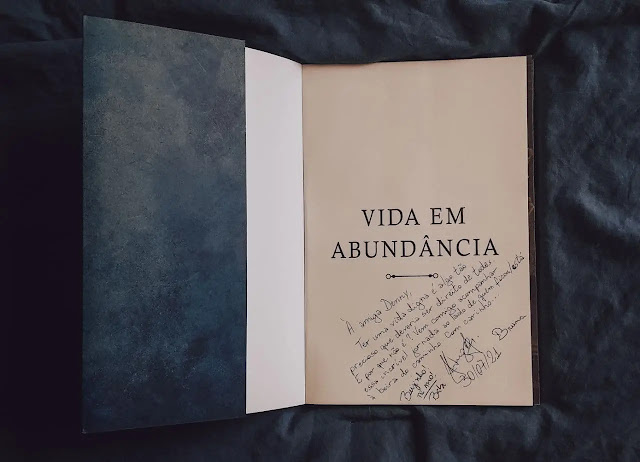 A imagem traz o livro aberto na primeira página. Nela, além do título, há o autógrafo do autor. Está escrito: "À amiga Denny. Ter uma vida digna é algo tão precioso que deveria ser direito de todes.  E porque não é? Vem comigo acompanhar essa incrível jornada ao lado de quemficou / está à beira do caminho. Com carinho..."  E então há sua assinatura,de sua esposa Babi e de sua filhinha Bruna.