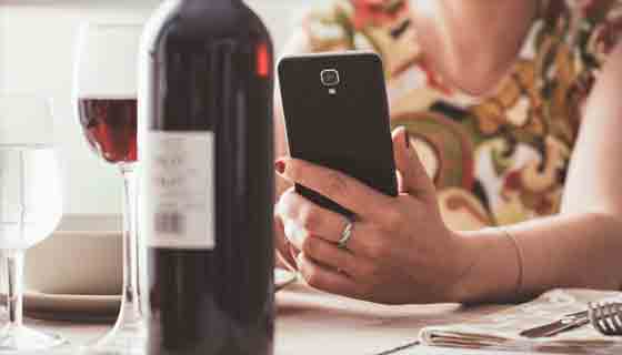 امرأة تتناول الغداء في المطعم وتستخدم تطبيق النبيذ مع هاتفها الذكي ، وهي تقوم بمسح ملصق زجاجة النبيذ