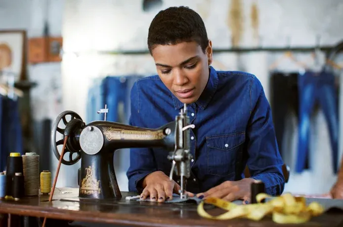 Person seated at sewing machine on table, holding and stitching denim piece, with thread spools and measuring tape nearby