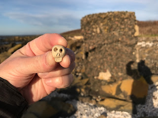 A photo showing a small ceramic skull (Skulferatu #65) being held up in front of the last of the anti tank blocks at Gosford Bay.  Photo by Kevin Nosferatu for the Skulferatu Project.