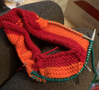 In progress: a piece of knitting with garter stitch and stockinette sections, with a stripe of red that is 3 ridges of garter stitch high, and a stripe of orange the same height. There are some green stitches on the needle but most are still orange.