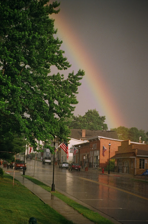 A rain slicked street going past some old red brick single story business buildings, with a rainbow arcing across the sky and ending above those buildings.