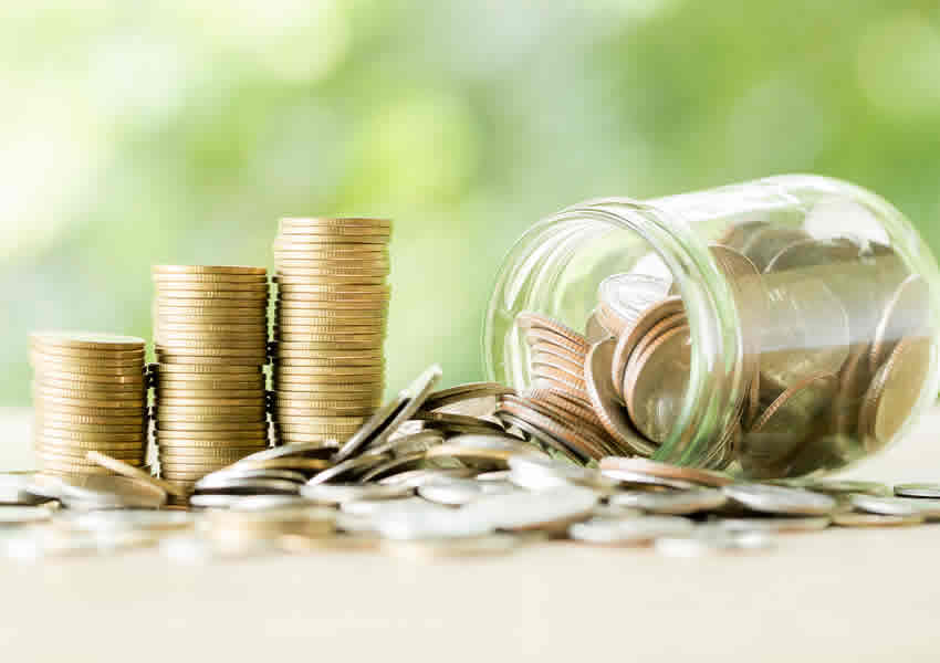 Imagem mostra 3 pilhas de moedas perto de um pode de vidro caído cheio de moedas com a tampa aberta e algumas moedas espalhadas.