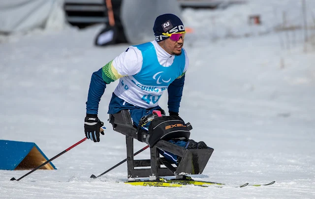 Cristian esqui usando um par de esquis adaptados. O brasileiro veste um agasalho azul e branco, com um colete com o número 40 por cima, e uma touca na cabeça