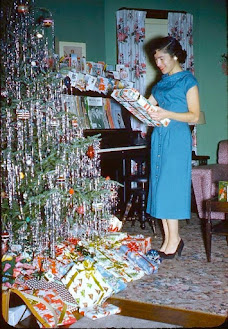 A photo from the 50s - it shows a woman in a blue dress and black heels in front of a heavily itself tree with lots of presents under it.