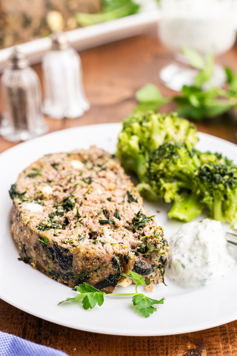 Slice of keto Greek style meatloaf of a white plate with broccoli and mashed cauliflower.