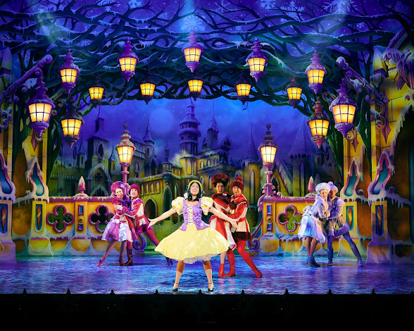 Snow White on stage in full voice, wearing the traditional costume, with three couples dancing behind her in different coloured flamboyant outfits