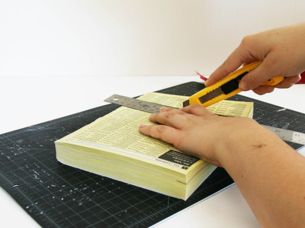 hands using craft knife and metal ruler to cut through phone book placed on cutting mat