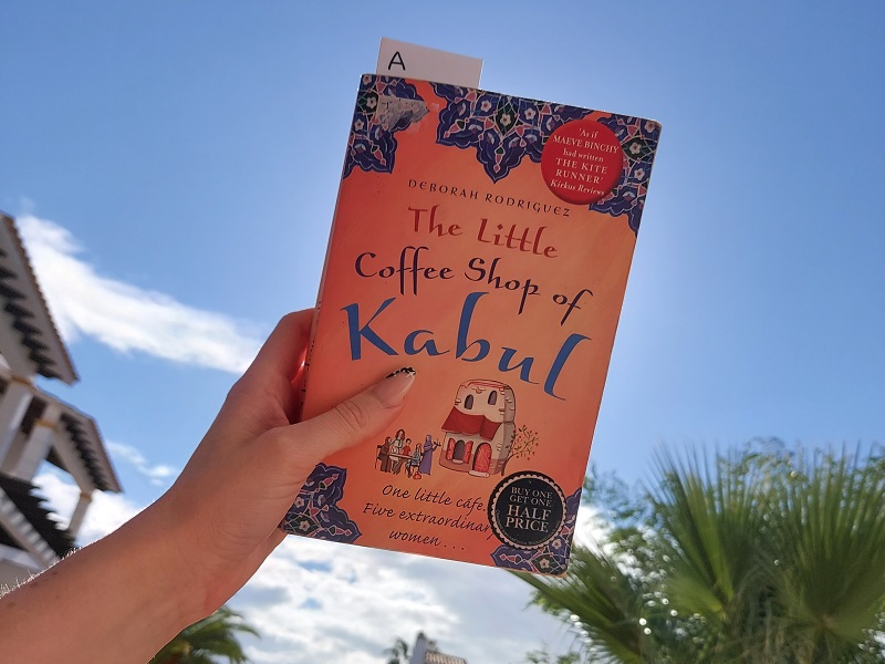 The Little Coffee Shop of Kabul book held against a blue sky, with palm trees and Spanish villas in background