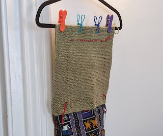 Photograph of a sagey-green garter stitch swatch marked with red yarn in a square and clipped to a coat hanger. A piece of multi colored black and purple fabric is suspended below