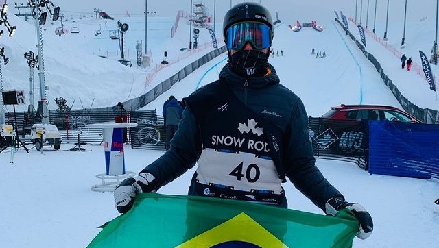 Sebastian Bowler posa para foto com a bandeira do brasil com uma montanha com neve no fundo