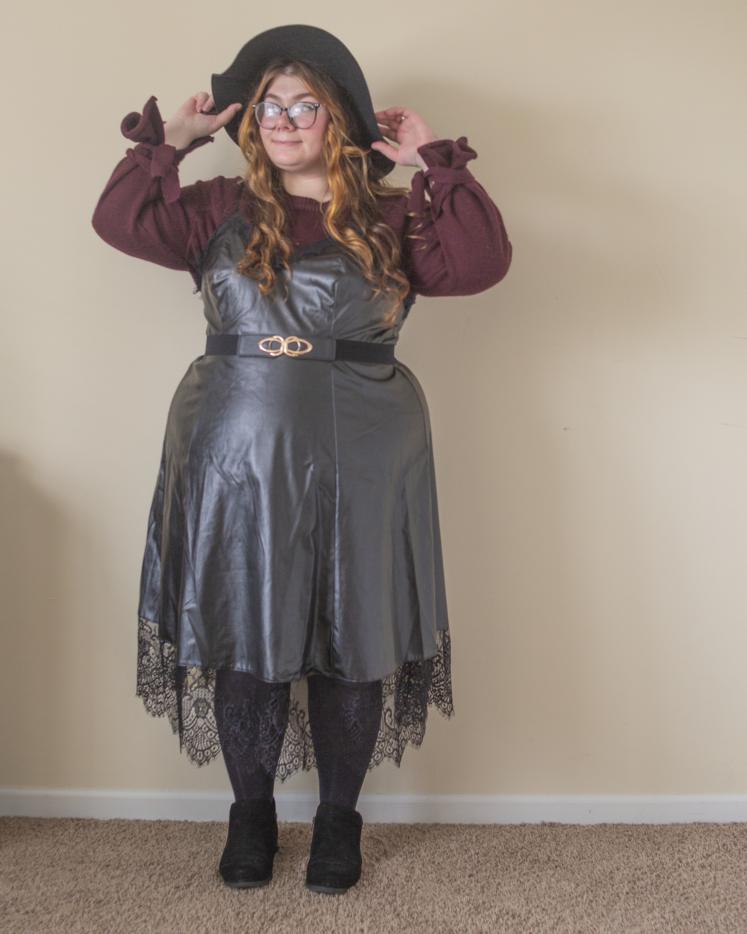 An outfit consisting of a berry knit sweater with ties around the wrists under a black leather midi dress with black lace trim, black tights and black boots.