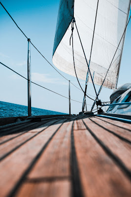 Dark wooden sailboat deck in forefront with white sail in background against a blue sky