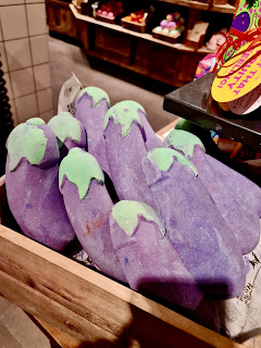 A photo of a bunch of large 10 inch purple aubergine shaped bath bombs with green and yellow tops in a large light brown box with a rectangular black card that says big aubergine in white font on a bright background