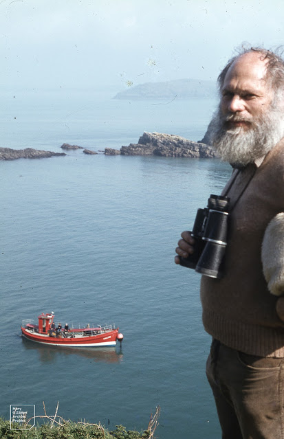 Bearded man with binoculars stands in front of the sea on a sunny day. In the background is a red, blue and white tour boat.