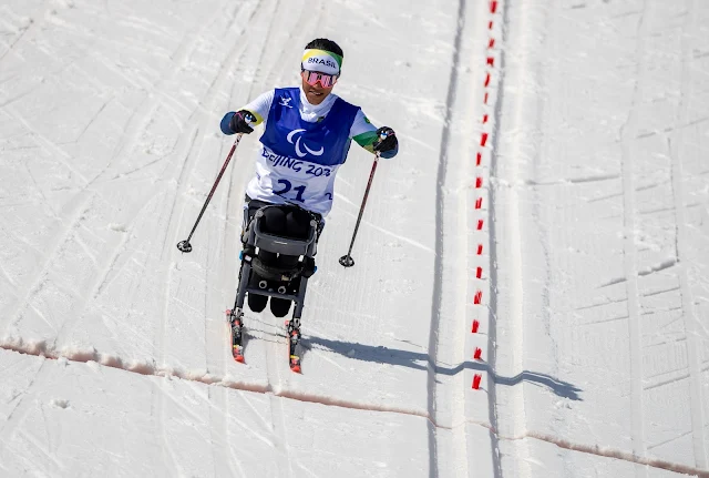 Aline esquia sentada usando esquis adaptados. Ela está no meio da neve usando um uniforme azul e branco e um colete com o número 21