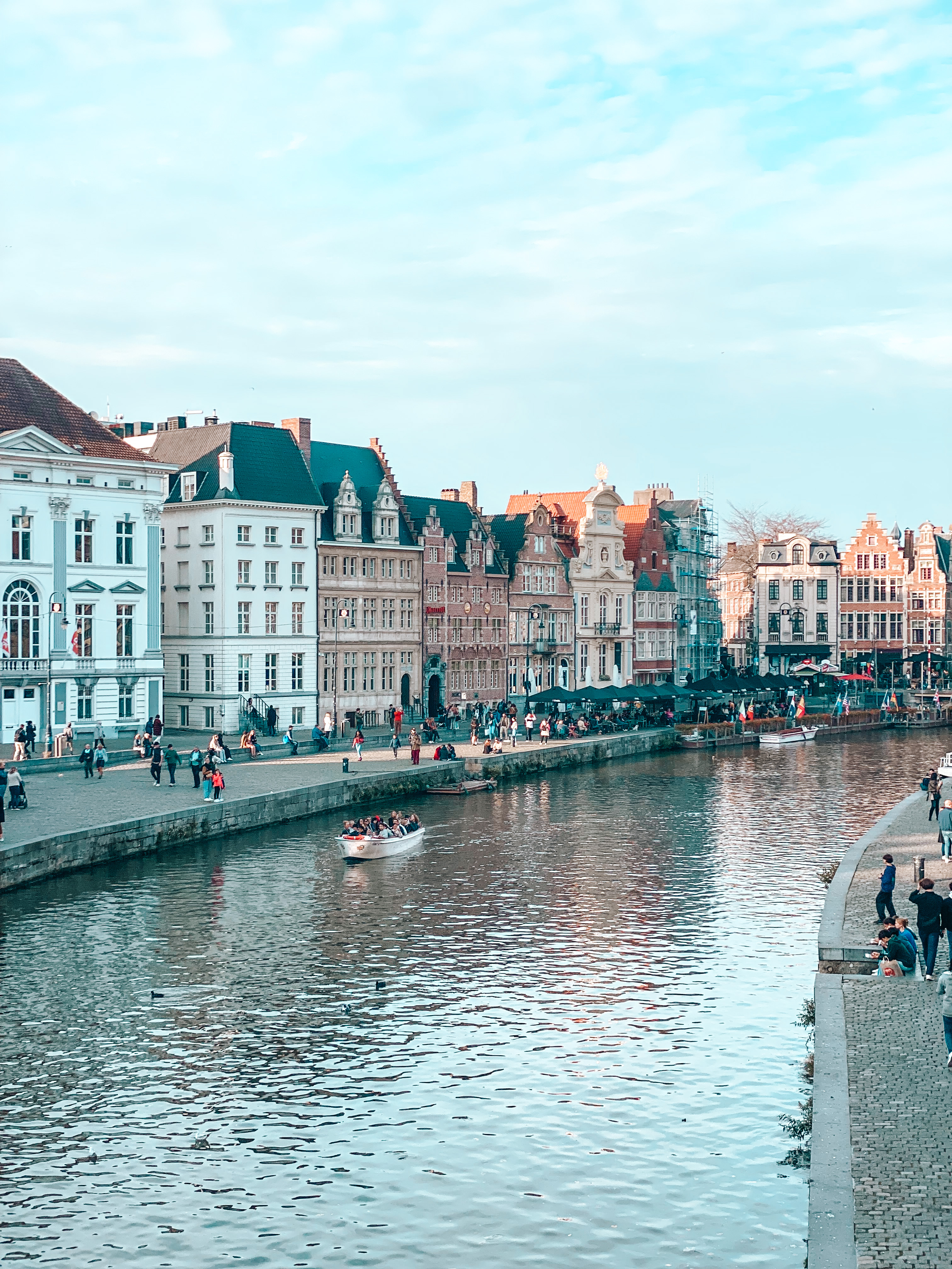 le long des canaux de gand les bâtiments de la ville à gauche et à droite et le fleuve au milieu avec un bateau qui s'y balade