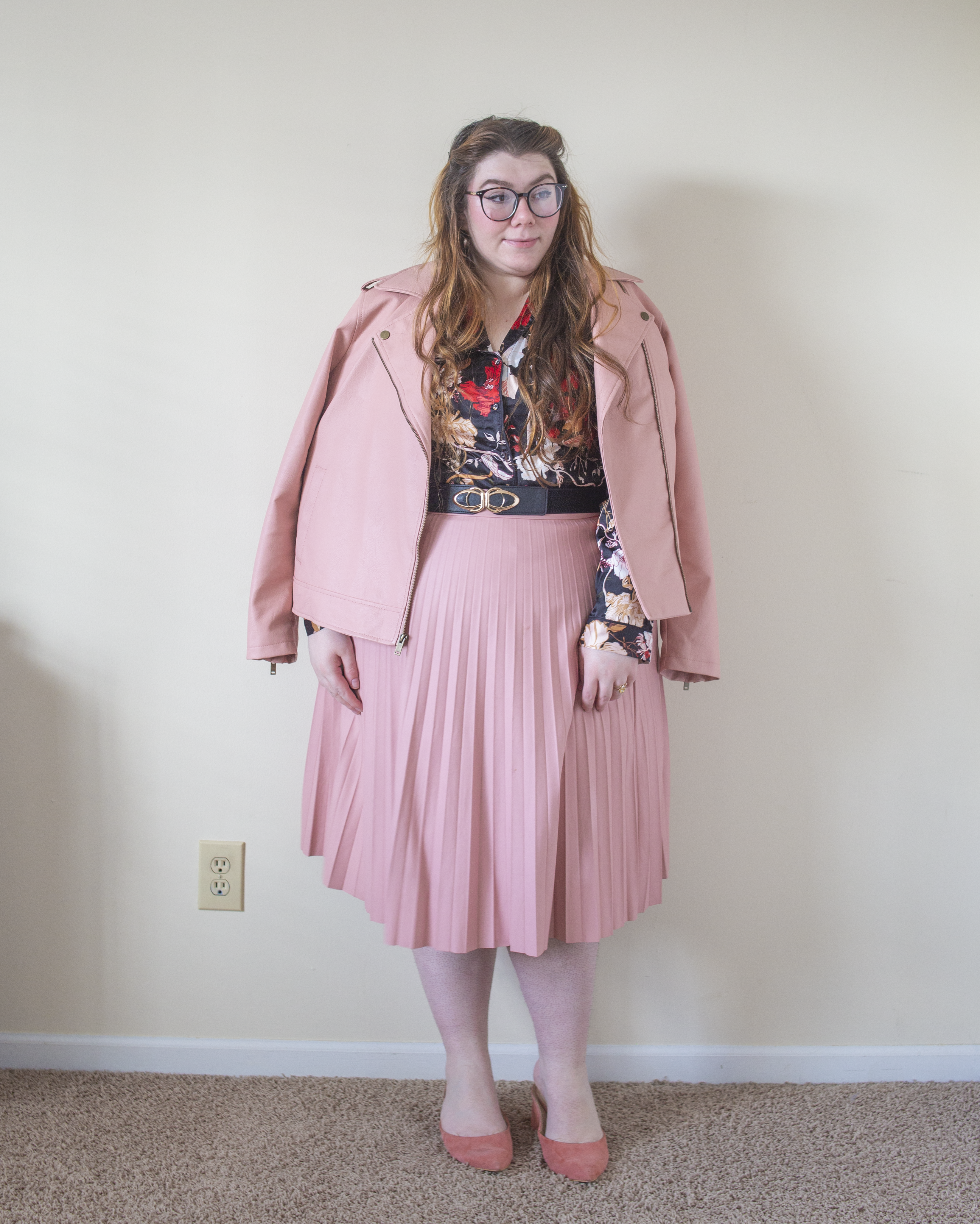 An outfit consisting of a pastel pink faux leather jacket over a black, cream, pink, yellow and red pajama style satin top tucked into a pastel pink leather pleated midi skirt and pink heeled clogs.