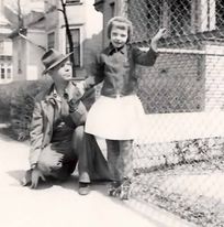 Dad kneeling, looking at, and holding the hand of a young girl who is on roller skates and clutching a chain link fence with the other her other hand