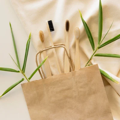 Multiple bamboo toothbrushes with colorful bristles displayed in brown paper bag surrounded by green plant leaves on cream background