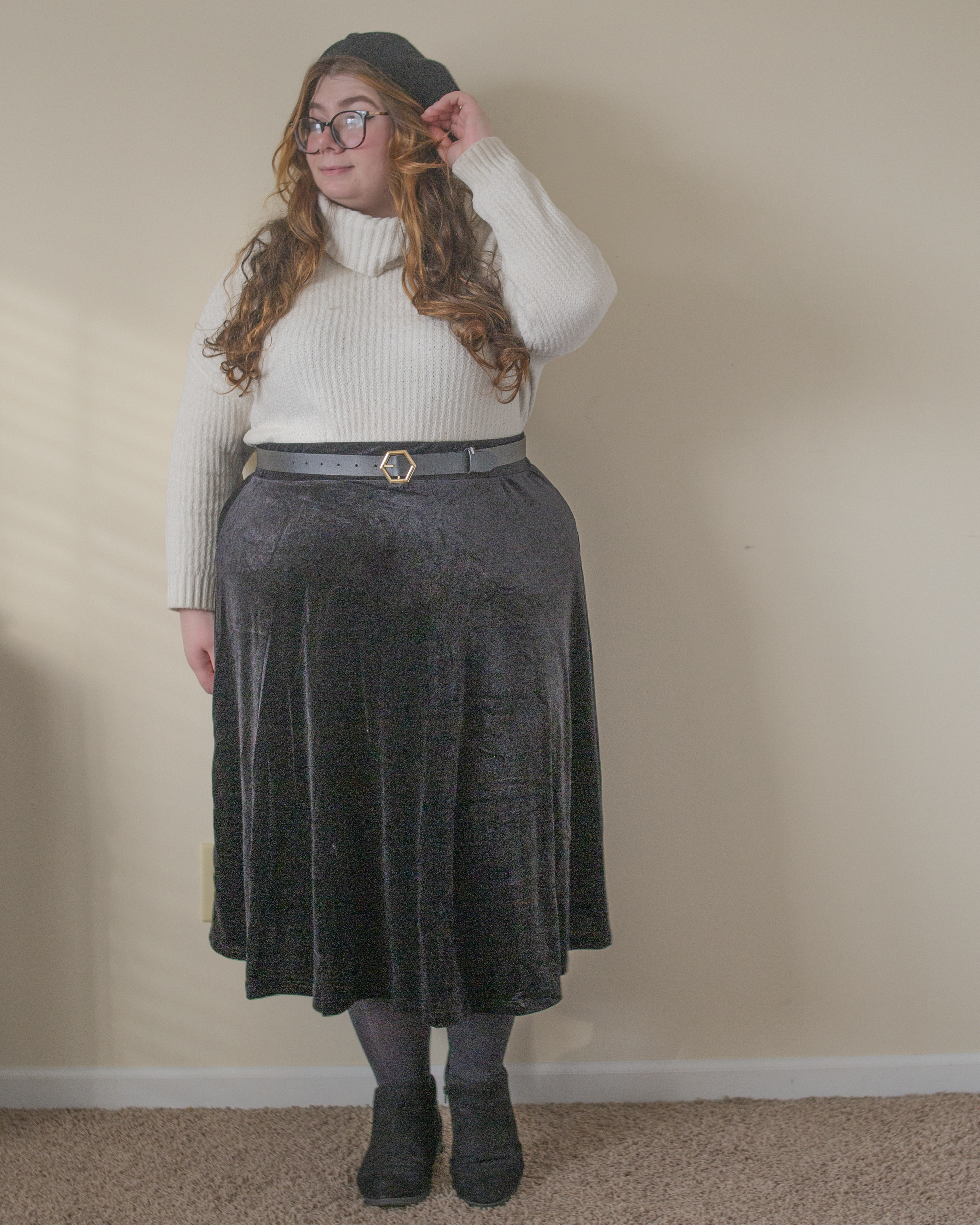 An outfit consisting of a white chenille turtleneck half tucked into a black velvet midi skater skirt, black tights, and black ankle strap heels.