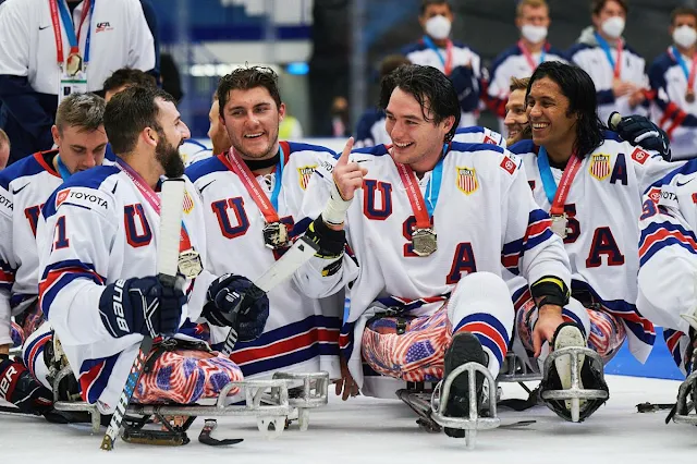 atletas dos Estados Unidos após receberem suas medalhas de ouro. Todos estão encima de seus trenós sorrindo e conversando. Eles vestem uma camisa branca escrito USA em azul na diagonal. Todos estão com uma medalha de ouro pendurada no pescoço