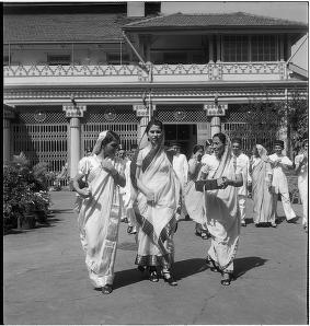 Três mulheres ao centro da fotografia caminham em direção à câmera. Ao fundo há mais pessoas e uma construção.