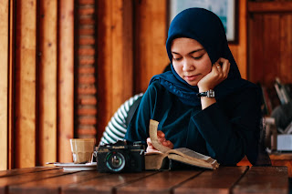 Image of Muslim female student in the library reading a book with a camera in front of her and a coffee drink beside her on the desk.