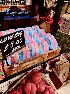 A Volkswagen Beetle shaped bath bomb with colour accents of blue, pink and red all over it with a black rectangular card that says love bug in white font next to it in a light brown rectangular box on a bright background